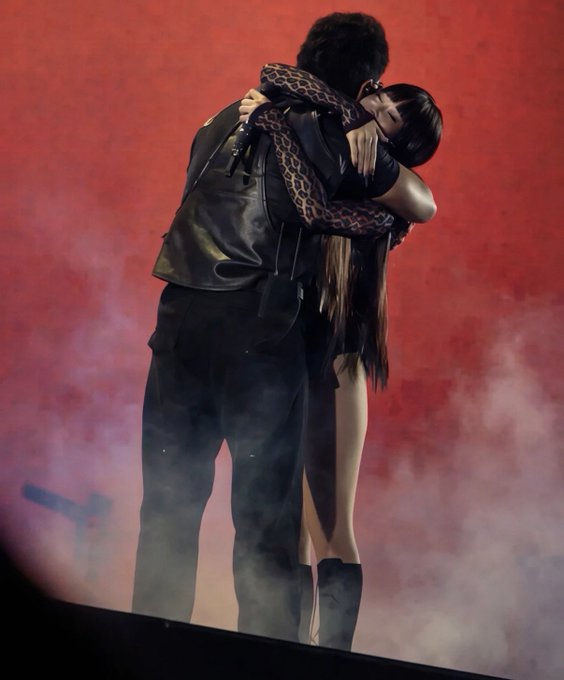 Two people embracing on a stage with a red background and smoke effects; one wears a black leather jacket and pants, while the other wears a black dress.