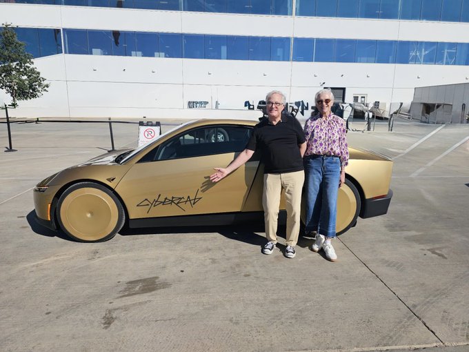 An older man and woman pose next to a gold Tesla Cybercab with "CYBER" graffiti on the side, parked in front of a modern glass building.
