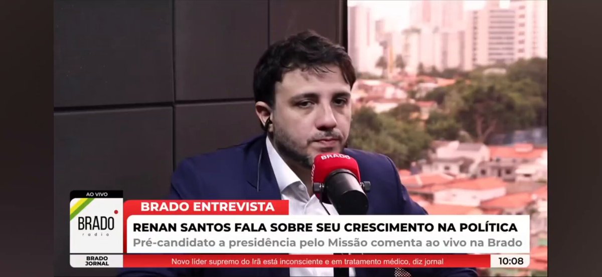 The image shows a man wearing a suit talking into a microphone at a news conference or event. The background of the image features a cityscape with tall buildings, suggesting that this could be taking place in a major urban area. The text on the image reads "BRADO ENTERTEVITA", which is likely the name of the news organization hosting the event.