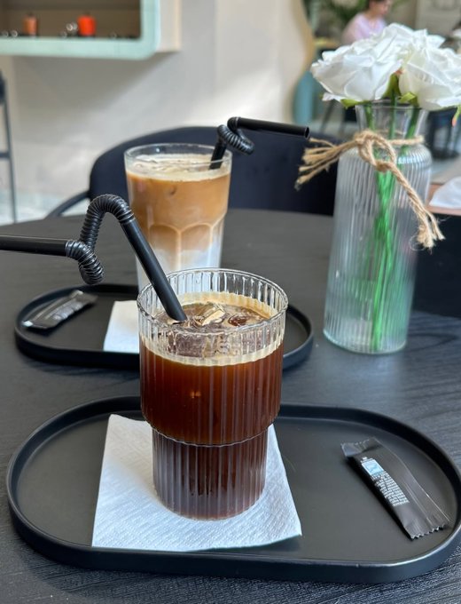 Two iced coffee drinks with black curly straws served on black trays at a cafe, with a vase of white roses tied with twine in the background.