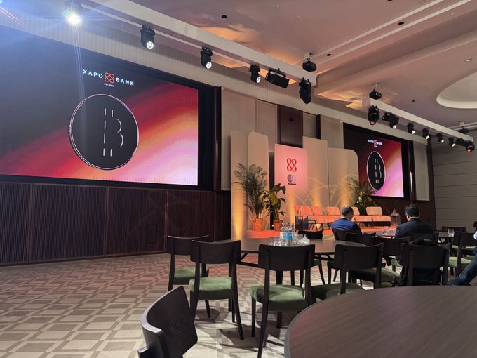 An interior view of a conference or event space with round tables, chairs, and large LED screens displaying the "XAPO BANK" logo and a Bitcoin symbol.