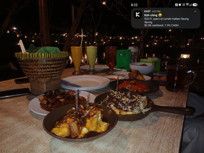 A nighttime dining photo showing various dishes on a table with a notification from KAST displaying "Kah-ching... $25.11 spent at rumah makan Saung Apung, $0.38 cashback - 1.5% CASH" at 8:22 PM.