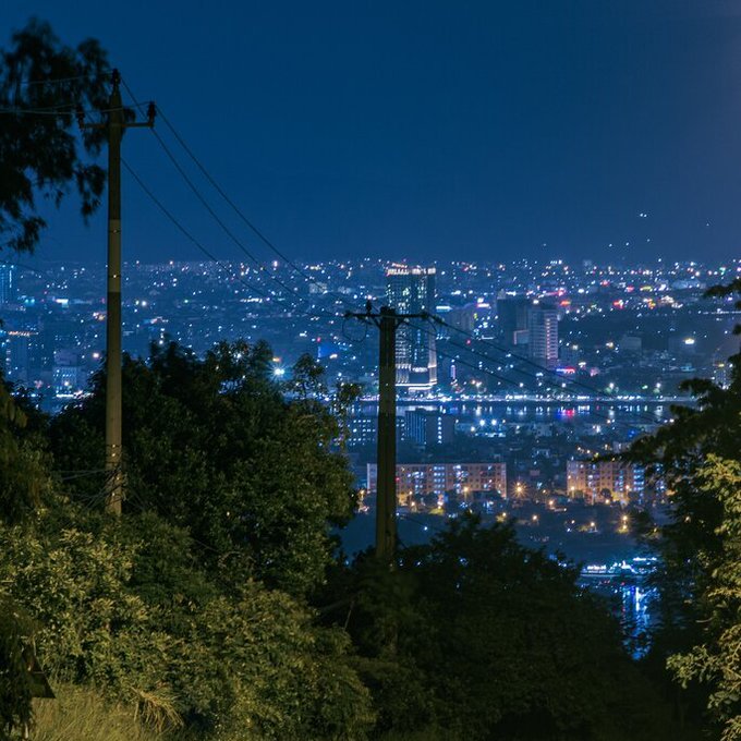 A nighttime cityscape view from an elevated position, framed by silhouetted trees and utility poles in the foreground.