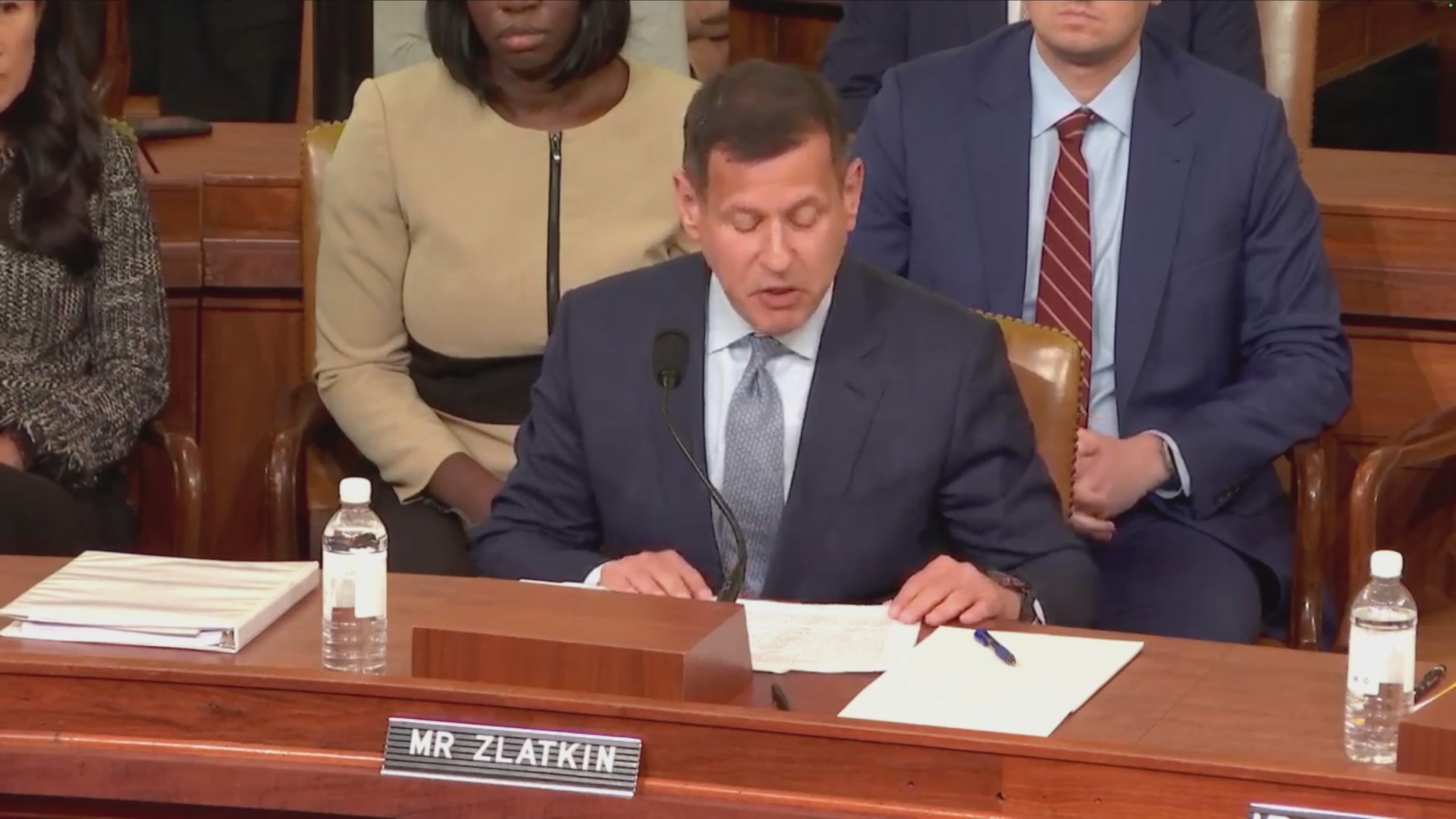 The image shows a man sitting at a wooden desk with papers in front of him while he speaks into a microphone during a hearing. The man appears to be dressed in a suit, and there are two other people present in the room who seem to be listening intently as he delivers his testimony. A water bottle is also visible on the table next to one of the individuals.