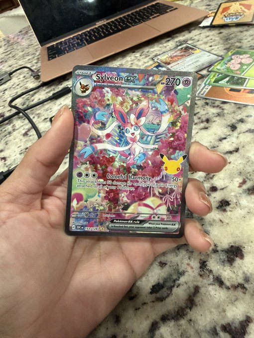 A hand holding a holographic Pokémon trading card featuring Sylveon ex and Pikachu with the text "Colorful Harmony" and "270" HP, displayed over a granite countertop near a laptop.
