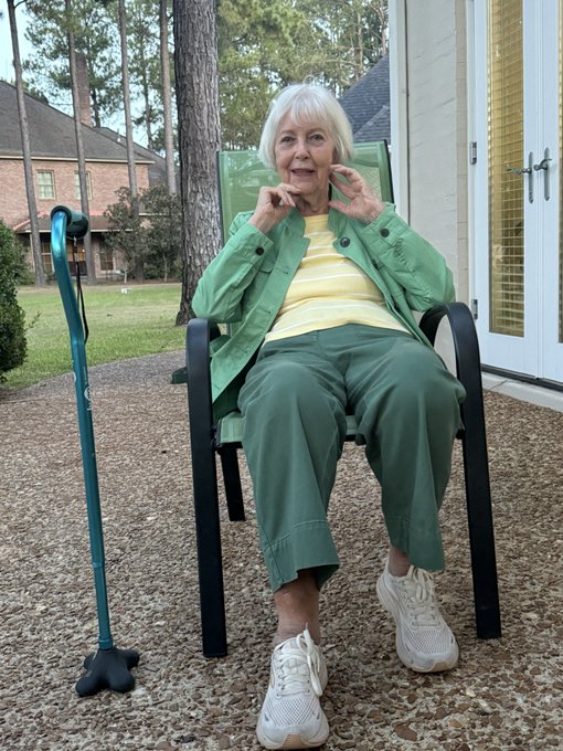 An elegant older woman with white hair sits in a green patio chair, wearing a mint green jacket over a yellow top with matching green pants and white sneakers, playfully framing her face with her hands as a teal cane rests beside her. The image perfectly embodies the "generational aura" mentioned in the caption—her confident pose, stylish coordination, and timeless elegance exude a charm that transcends age, proving that grace and presence are their own kind of inheritance.
