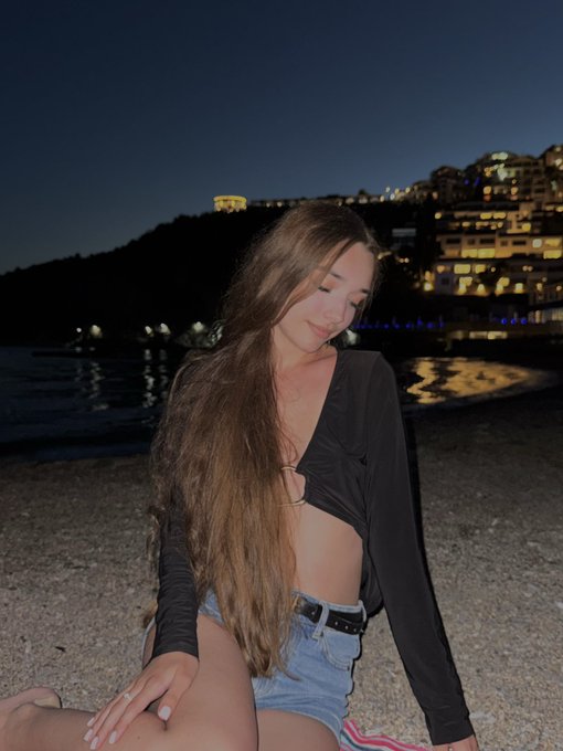 Photograph of a young woman with long brown hair sitting on a beach at night, wearing a black long-sleeve crop top and denim shorts, with illuminated buildings on a hillside in the background.