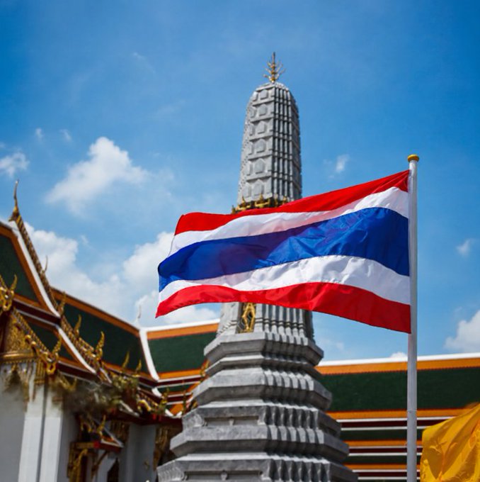 The Thai flag waves prominently in the foreground, with a tall ornate temple spire and traditional Thai architecture visible behind it against a blue sky with scattered clouds.