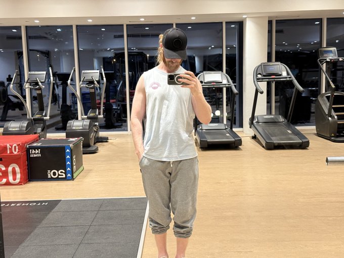 A man takes a mirror selfie in a gym, flexing slightly in a white tank top, gray shorts, and black cap. The caption plays on his relaxed physique and gym setting, jokingly whispering self-motivations to "be strong" for $JAMES and Monad while on the "road to 9 figures."