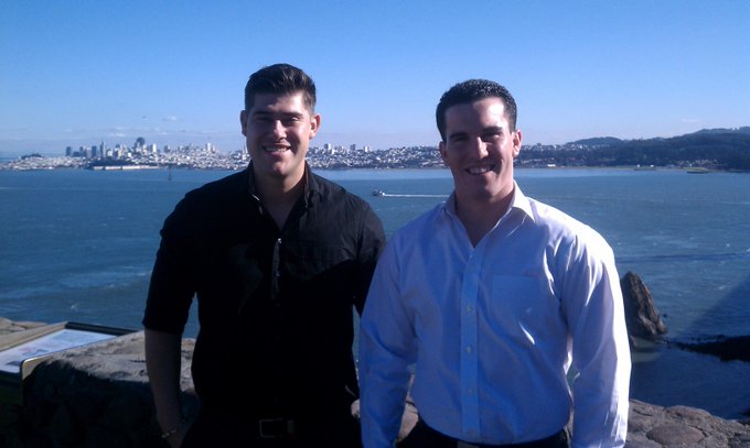 Two smiling men pose for a photo against a scenic backdrop of a bay with a city skyline visible across the water, suggesting a moment of celebration or milestone marking. The image accompanies a post celebrating 15 years of building KiiChain and its mission of creating real change, with the picturesque setting symbolizing the journey and future ahead.