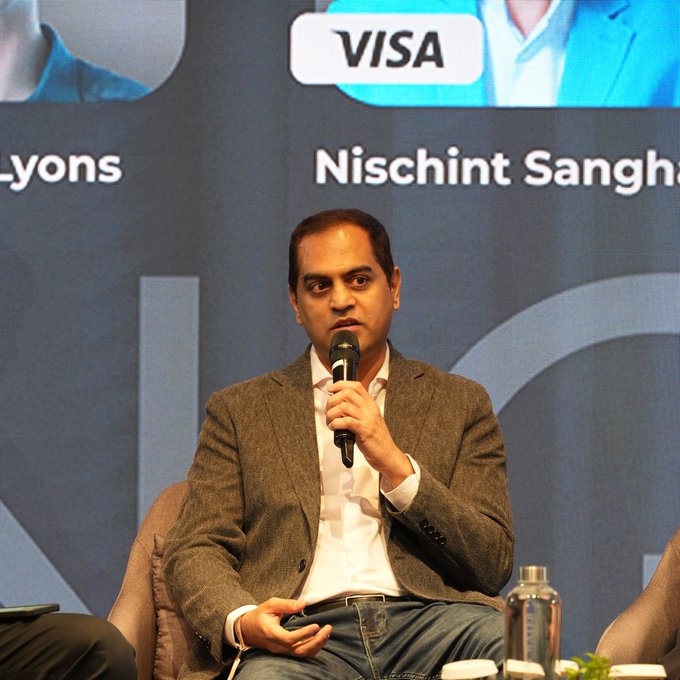 A man identified on screen as "Nischint Sangha[vi]" speaks into a microphone at a Visa-sponsored panel discussion. The image directly accompanies the quote from Visa's Head of Digital Currencies, visually representing the executive making the statement about the future of weekend payments becoming obsolete.