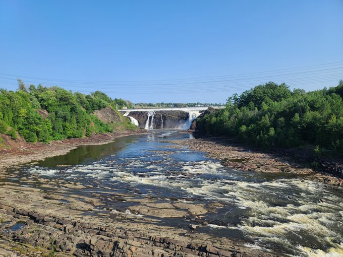 A scenic waterfall cascades over a concrete dam into a rocky river, surrounded by lush green trees under a clear blue sky. The "Gm frens" greeting pairs perfectly with the bright, refreshing morning vibes of this peaceful nature view.