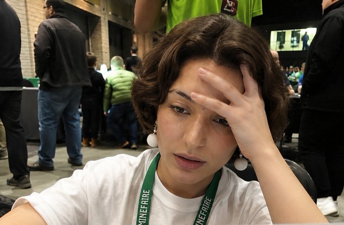 A person with short brown hair and a green 'MINEFAIRE' lanyard resting their hand on their forehead at what appears to be a convention or event.