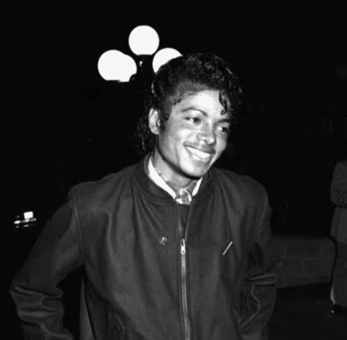 A black-and-white archival photograph of a smiling young Michael Jackson wearing a dark zip-up jacket, standing in front of an ornate lamppost at night.