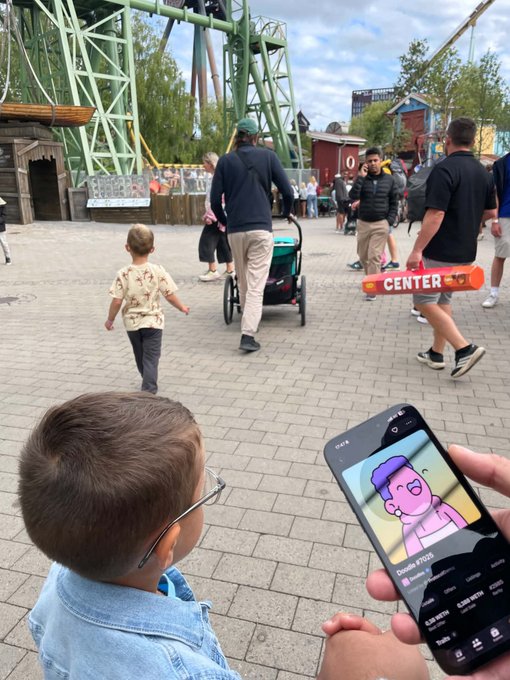 A young child wearing glasses watches a cartoon on a smartphone held by someone, while looking toward an amusement park with a roller coaster in the background.
