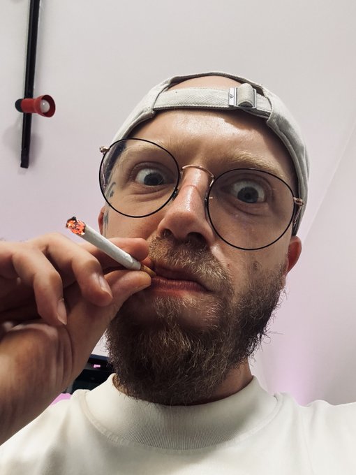 A man with a beard, glasses, and a backwards cap makes a wide-eyed expression while holding a lit cigarette near his mouth.