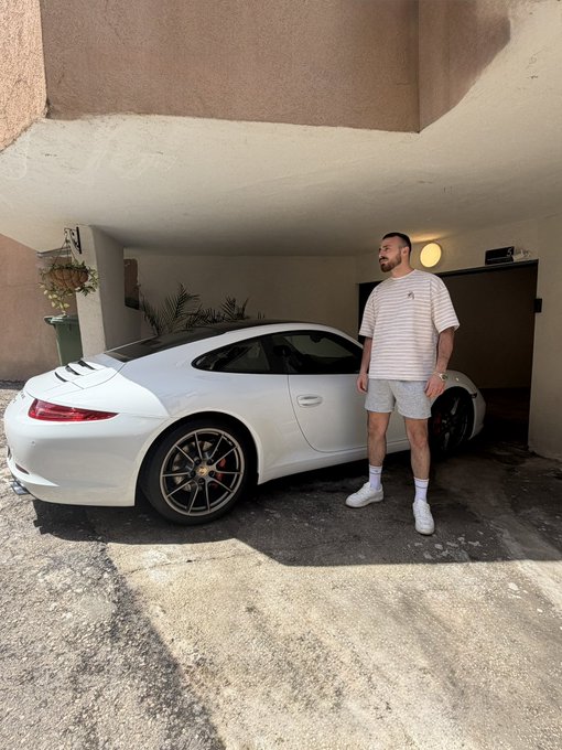 A man in a white striped shirt, gray shorts, and white sneakers stands proudly beside a white Porsche 911 in a covered parking area, sharing a cheerful "Gm friends" greeting with his followers.
