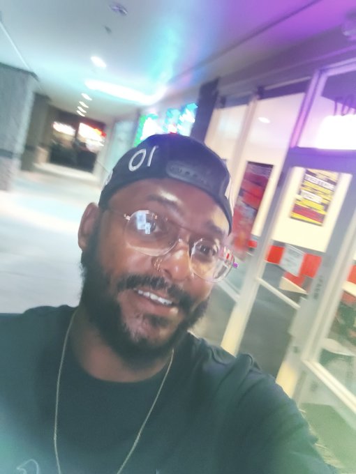 A selfie of a smiling bearded man wearing glasses, a backwards black cap with "01" on it, and a dark shirt, taken in what appears to be a parking garage.