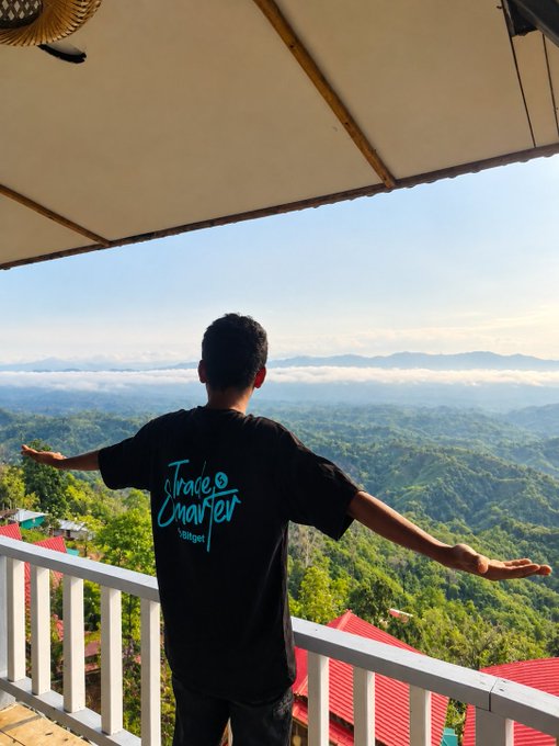 A man viewed from behind wearing a black 'Trade Chapter Vlogger' t-shirt stands on a balcony overlooking a mountainous landscape with his arms outstretched.