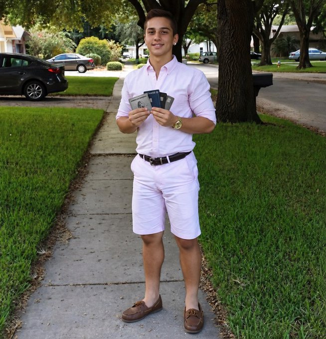 A young man wearing a pink button-down shirt, white shorts, and brown loafers stands on a suburban sidewalk holding a fan of cash, with cars and trees in the background.