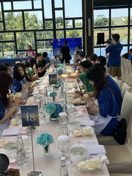 A group of students sitting at a long dining table labeled '18' in a bright room with large windows, participating in what looks like a school meal or event.