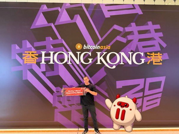 A man stands on stage at Bitcoin Asia holding a 'Reality Tokens Empowering Millions' sign next to a large white cartoon mascot, with 'HONG KONG' displayed on the screen behind him.