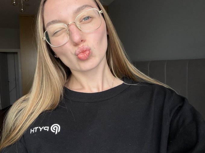 The image features a woman with long blonde hair wearing glasses, making a funny face as she poses for the camera. She has a black shirt on that says "world cup powered by @PythNetwork" written on it. The background of the photo is blurred and out of focus, drawing attention to the woman in the foreground.