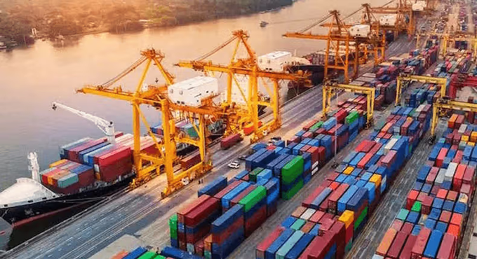 An aerial view of a busy shipping port at sunset, featuring large yellow gantry cranes loading colorful stacked shipping containers onto cargo vessels.