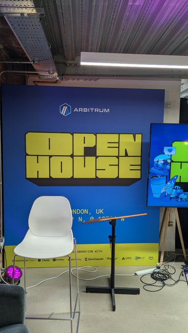 An 'Arbitrum Open House' event setup in London, UK, featuring a blue branded backdrop, a white chair, and a music stand.