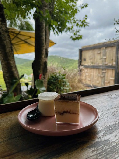 A plate holding a small jar and a dessert in a clear cup labeled 'Banh Minh Oi' sits on a wooden railing overlooking lush green mountains under a cloudy sky, with a yellow umbrella to the side.