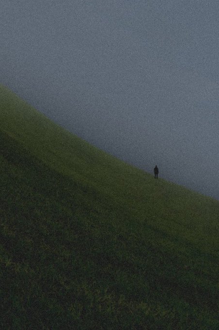 An aerial view showing a lone figure walking along the edge of a grassy hillside meeting a gray paved surface, creating a stark diagonal composition.