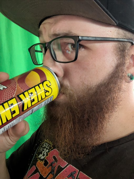 A close-up of a bearded man wearing glasses and a cap, drinking from a yellow and red beverage can, against a bright green background.