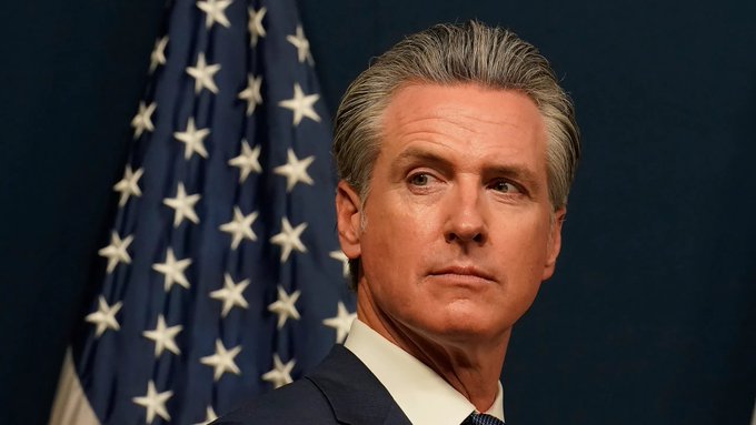 California Governor Gavin Newsom stands in front of an American flag, looking serious. The image accompanies news of his initiative to ban public officials from launching memecoins.