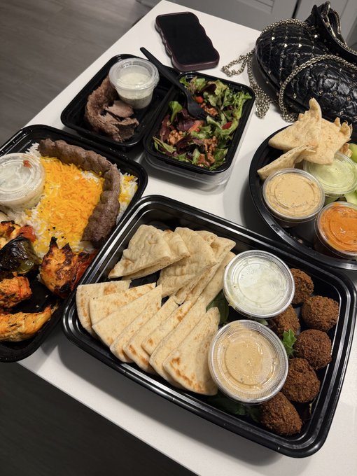 An overhead shot of a takeout meal spread on a white table, featuring multiple black plastic containers with pita bread, falafel, kebabs, rice with saffron, salad, and various dipping sauces, alongside a black quilted purse.