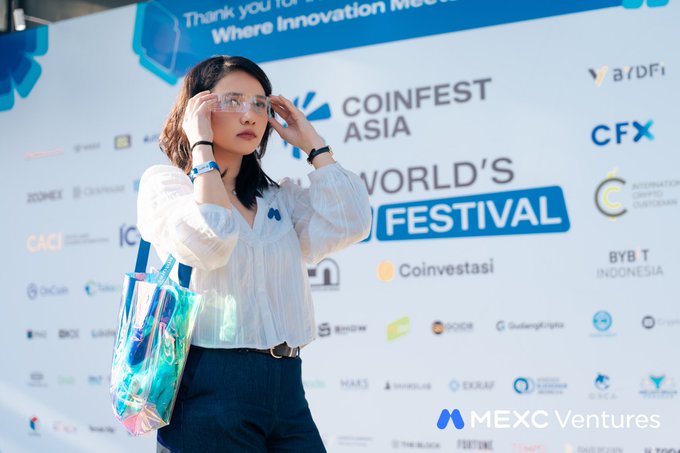 A woman with clear glasses stands in front of a Coinfest Asia backdrop featuring logos for MEXC Ventures, BYB Indonesia, and 'World's Festival'.