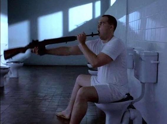 The image shows a man sitting on a toilet with a gun held above his head. The scene appears to be set in an indoor bathroom, possibly a public restroom or a hotel room. There are two toilets visible in the image - one being used by the man holding the gun and another located further away from him. The post about Bitcoin falling below $60,000 is unrelated to the image and does not provide any context for its relevance.