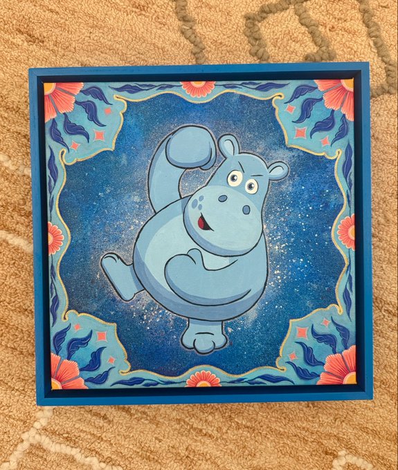 A small square painting of a cartoonish blue hippo with a cheerful expression, set against a starry dark blue background with decorative floral and leaf motifs in the corners, framed in blue.