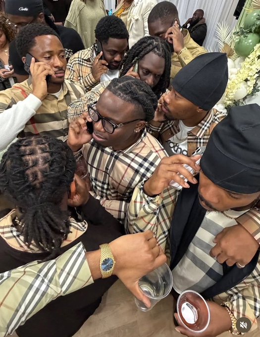A group of men wearing matching Burberry-patterned shirts and outfits are gathered closely together, with some talking on phones and one wearing a black cap.