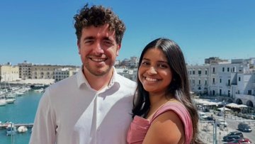 A couple smiling for a photo outdoors, with the man in a white shirt and the woman in a pink top, set against a sunny coastal town with white buildings and a harbor.
