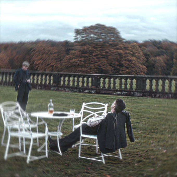 An atmospheric photo of a man relaxing in a chair on a lawn overlooking a stone balustrade, with another figure standing in the background near autumn trees.