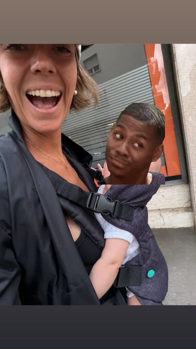 A smiling woman wearing a black jacket and a baby carrier, with the man visible in the background.