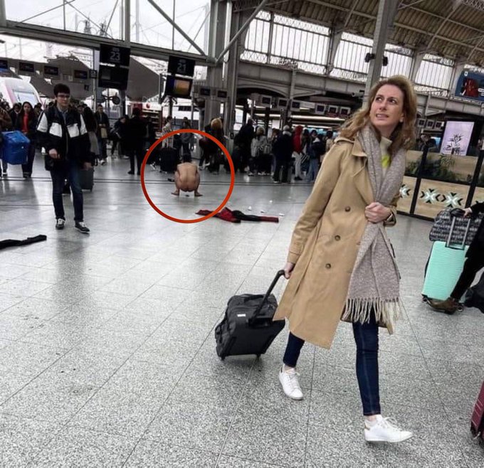 A woman in a tan trench coat and scarf stands calmly with her rolling suitcase in a busy train station, while a bold man in red shorts (circled) performs a daring handstand in the middle of the concourse behind him. Platform "25" is visible in the background, capturing the playful contrast between everyday commuters and someone taking a spontaneous leap into the spotlight.