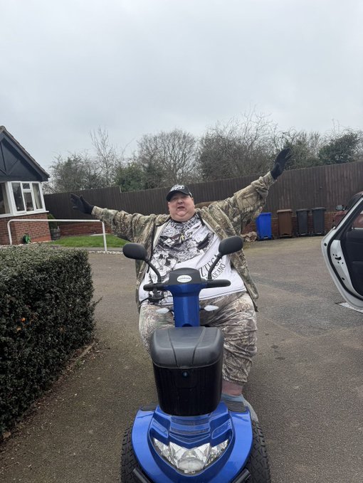 A man in a camouflage jacket, camo shorts, and a black cap sits on a blue mobility scooter with his arms outstretched in a residential driveway.