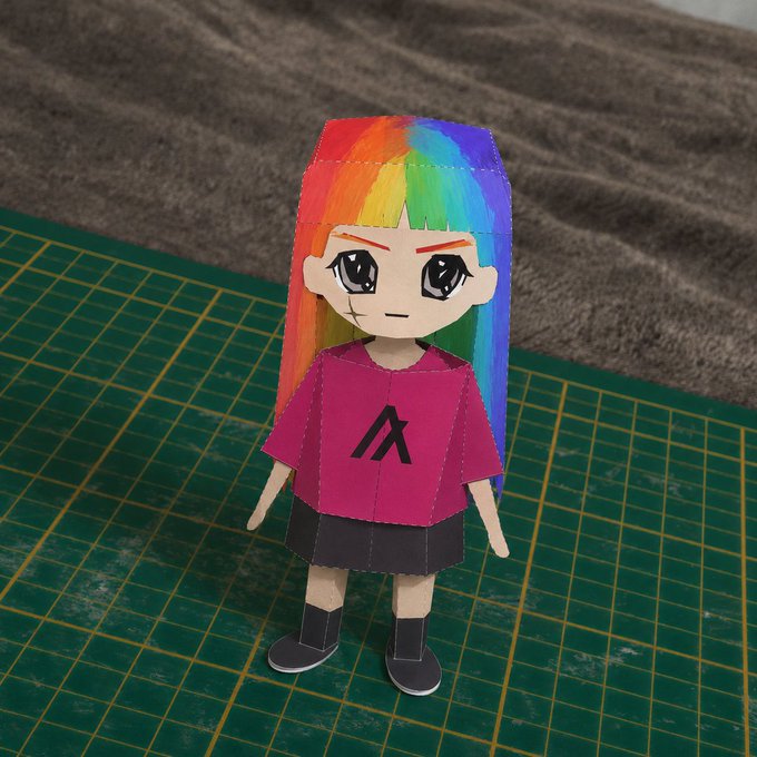Handmade papercraft figure of a character with rainbow-colored hair, an angry expression, wearing a magenta shirt with an 'A' logo and a black skirt, displayed on a green cutting mat.