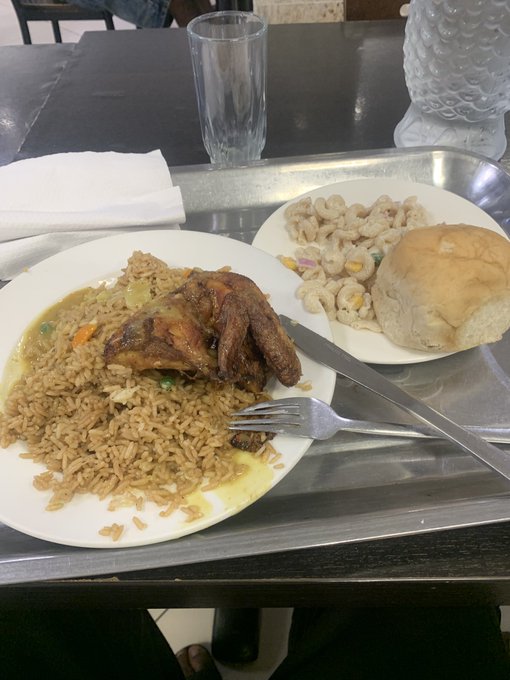 A metal cafeteria tray holds a plate of rice with chicken and gravy, a fork and knife, and a side plate with macaroni and cheese and a dinner roll, with an empty drinking glass in the background.