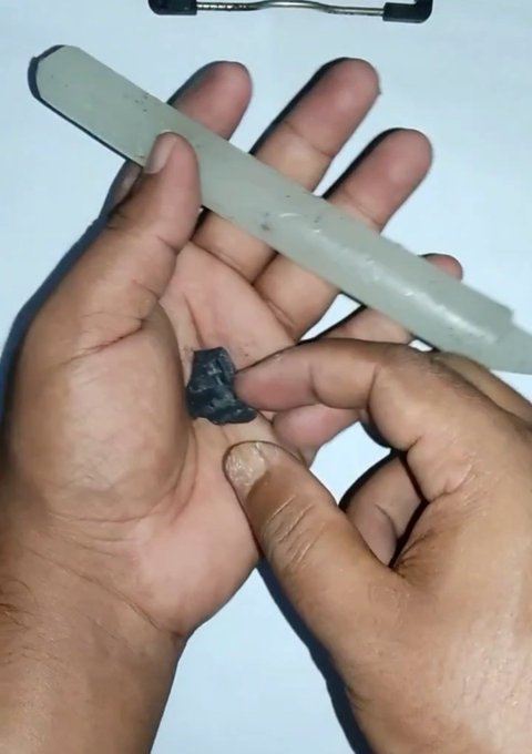 A close-up photo of two hands, one holding a small dark object (possibly a stone or piece of hardware) while the other holds what appears to be a metal tool or blade above it.