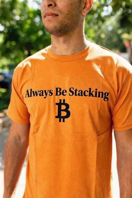 A man wearing an orange t-shirt with the text 'Always Be Stacking' printed in black above a Bitcoin (₿) symbol, standing outdoors with greenery in the background.