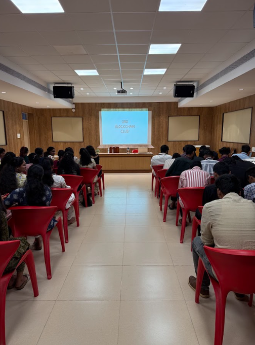 A conference room with attendees seated in red plastic chairs facing a projection screen displaying 'GFB Blockchain Club' on a wooden-paneled stage.