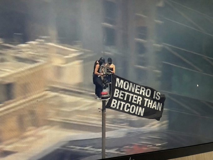 A photograph of two people on a motorcycle holding a banner that reads 'MONERO IS BETTER THAN BITCOIN' in an urban setting.