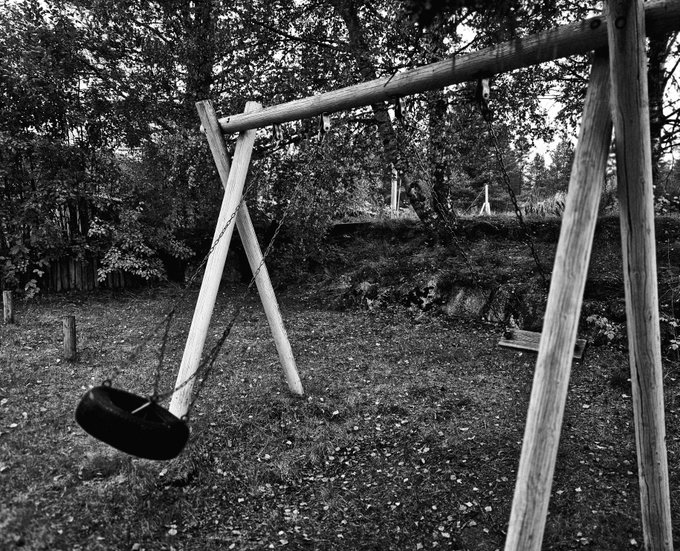 A black and white photograph of a wooden swing set with a tire swing hanging from chains in a grassy, leaf-strewn yard surrounded by trees.